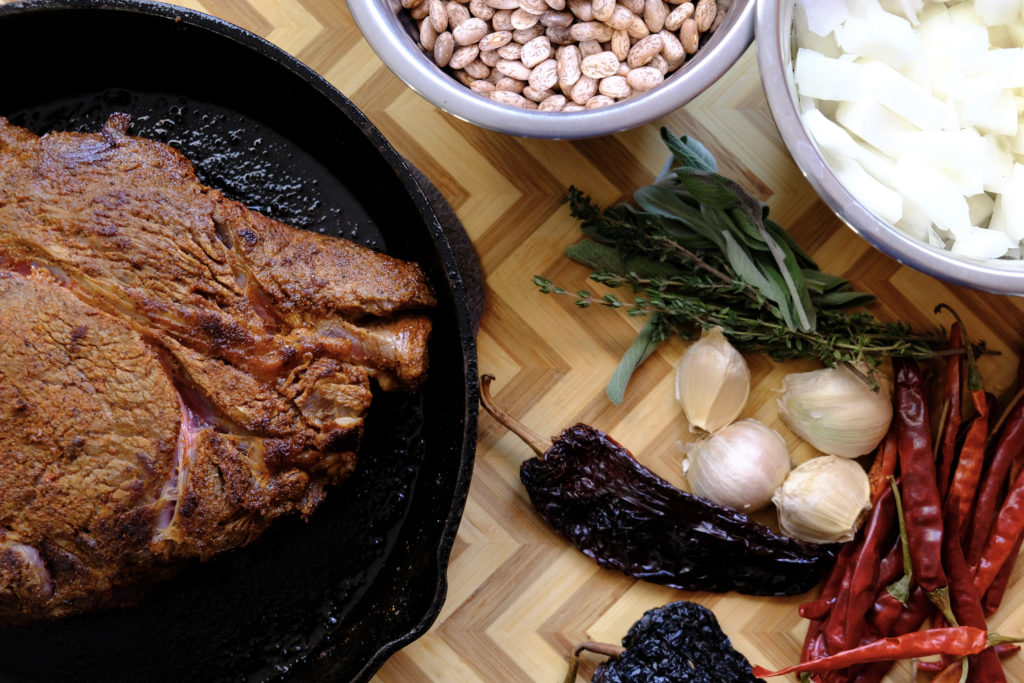 A beef chuck roast in a cast iron pan, surrounded by New Mexico ingredients like beans, garlic, and peppers
