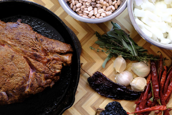 A beef chuck roast in a cast iron pan, surrounded by New Mexico ingredients like beans, garlic, and peppers